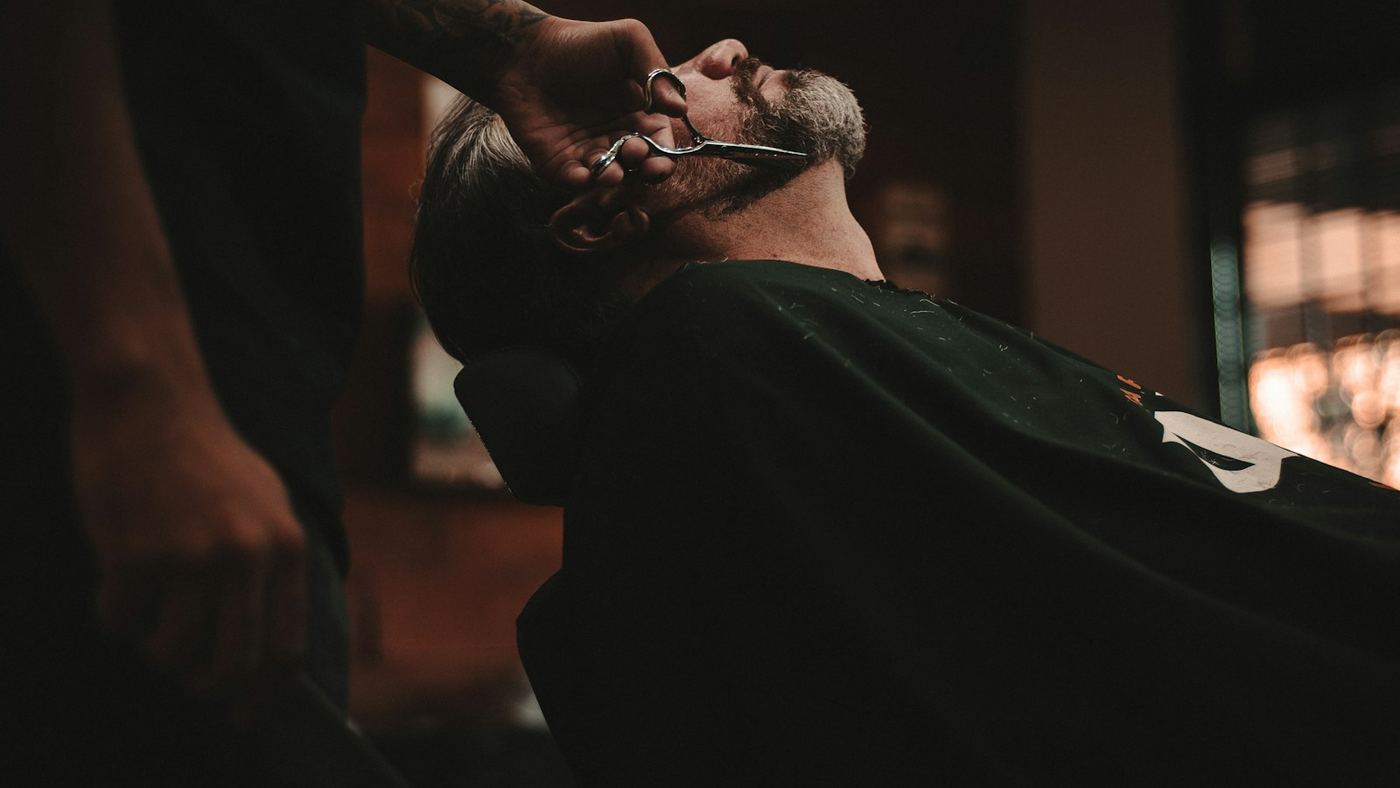 A barber giving a client a straight-razor shave in a darkly lit barbershop